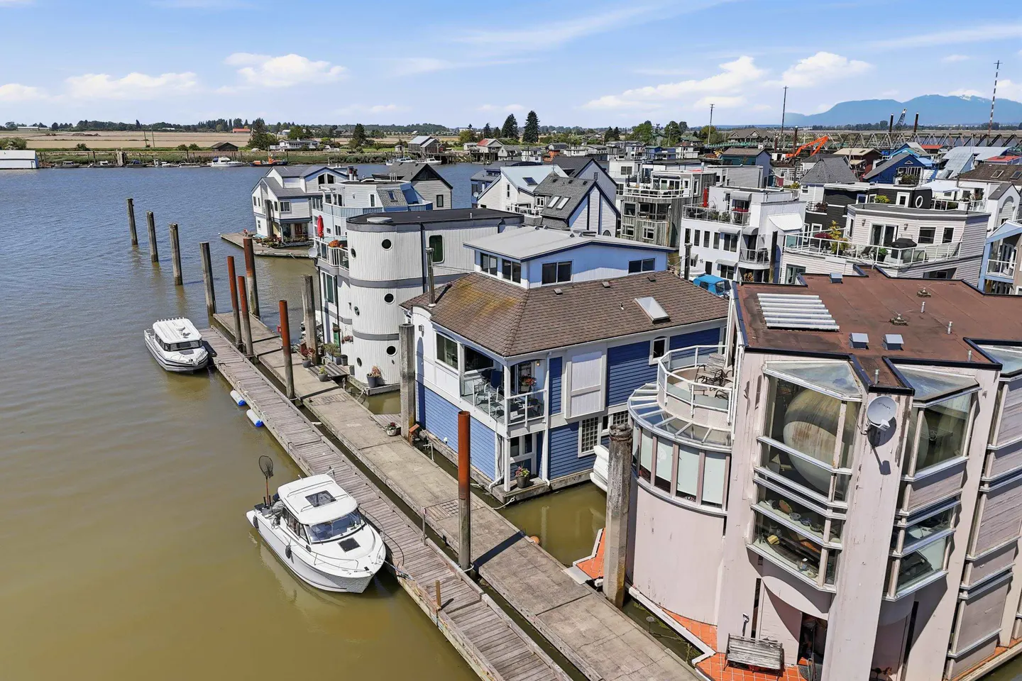 Aerial view of a houseboat community on a river, with boats docked at wooden piers. Houses are various colors and styles.