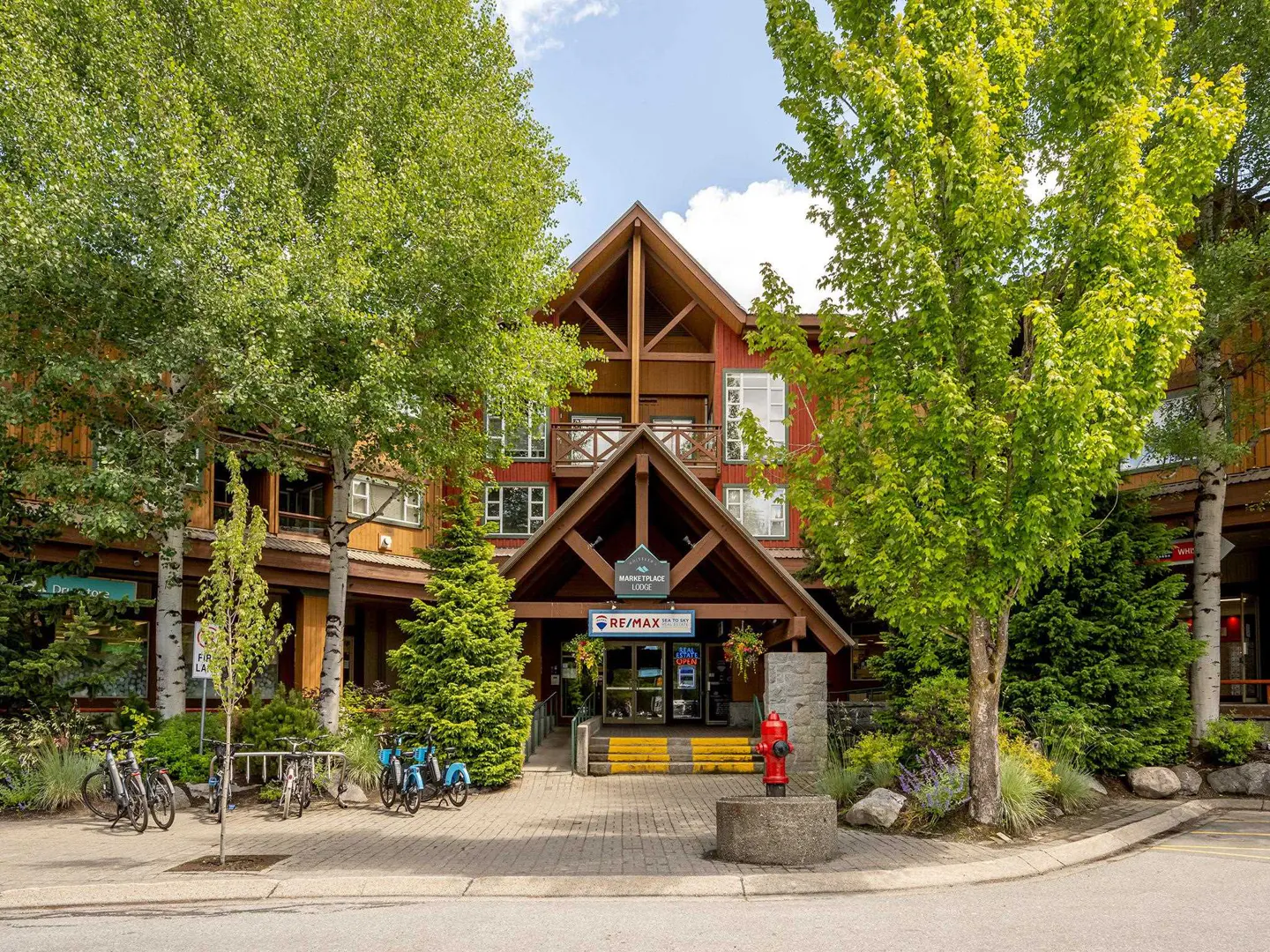 Exterior of Marketplace Lodge, a red and brown building with a peaked roof, surrounded by green trees and parked bicycles. A RE/MAX sign is visible.
