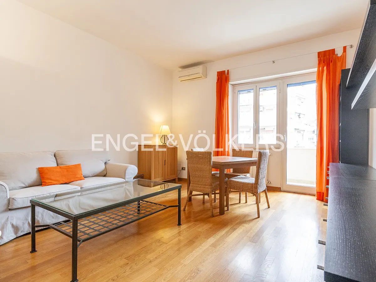 Living room with hardwood floors, a beige sofa with an orange pillow, a glass coffee table, and a dining table with four wicker chairs. Orange curtains frame a balcony door.