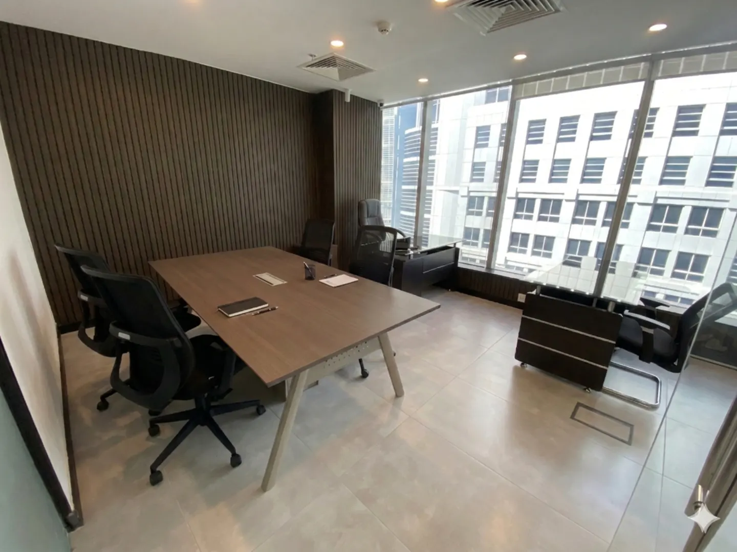 Bright office space with a large window view. A brown conference table is surrounded by black chairs.