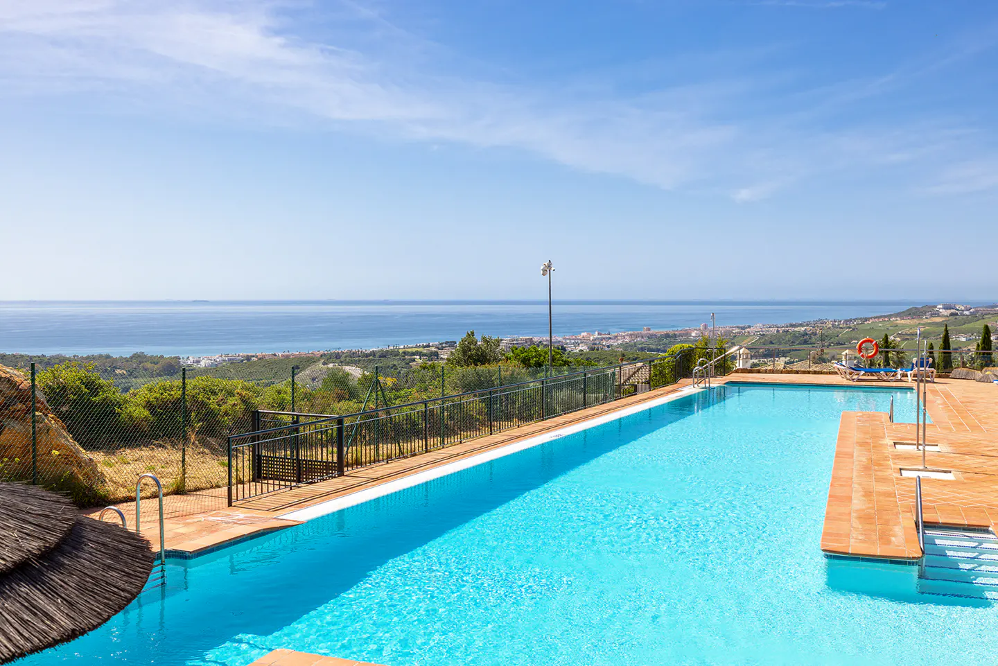 A long, blue swimming pool with a brick deck overlooks the ocean and a distant city under a clear, blue sky.