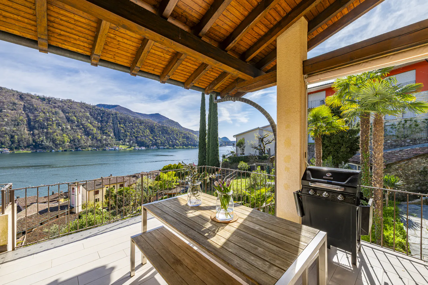 Covered patio with wood table, bench, grill, and lake view. Vases of flowers sit on the table.