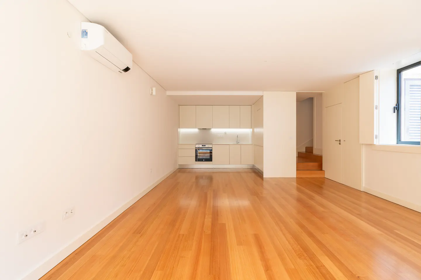 Bright, empty apartment with hardwood floors, white walls, and a compact kitchen area with light beige cabinets. A window and stairs are visible.