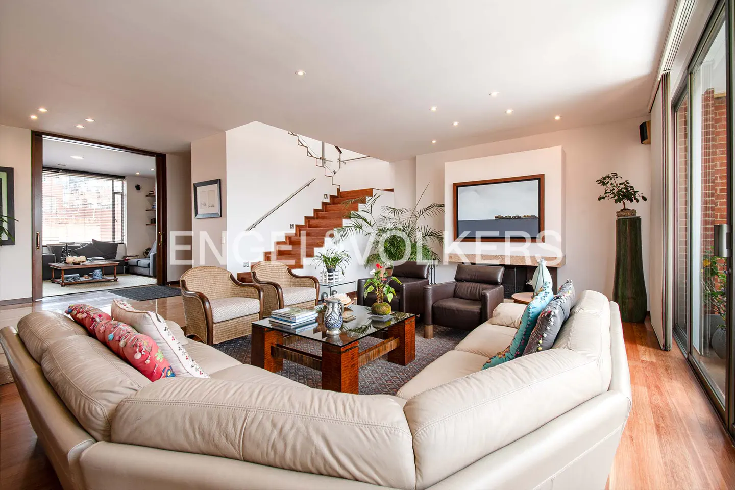 Bright living room with a beige sofa, wood floors, and a glass coffee table. A staircase and a fireplace add architectural interest.