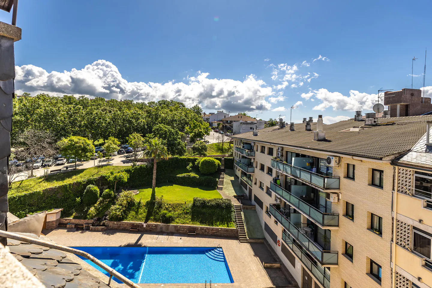 View of a blue swimming pool, green lawn, and apartment building under a blue, cloudy sky.