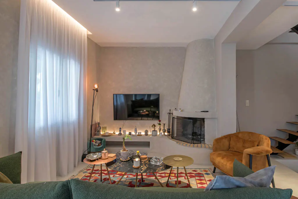Living room with a fireplace, TV, and seating. The walls are gray, and the floor is covered with a colorful rug. There are sheer white curtains on the window.