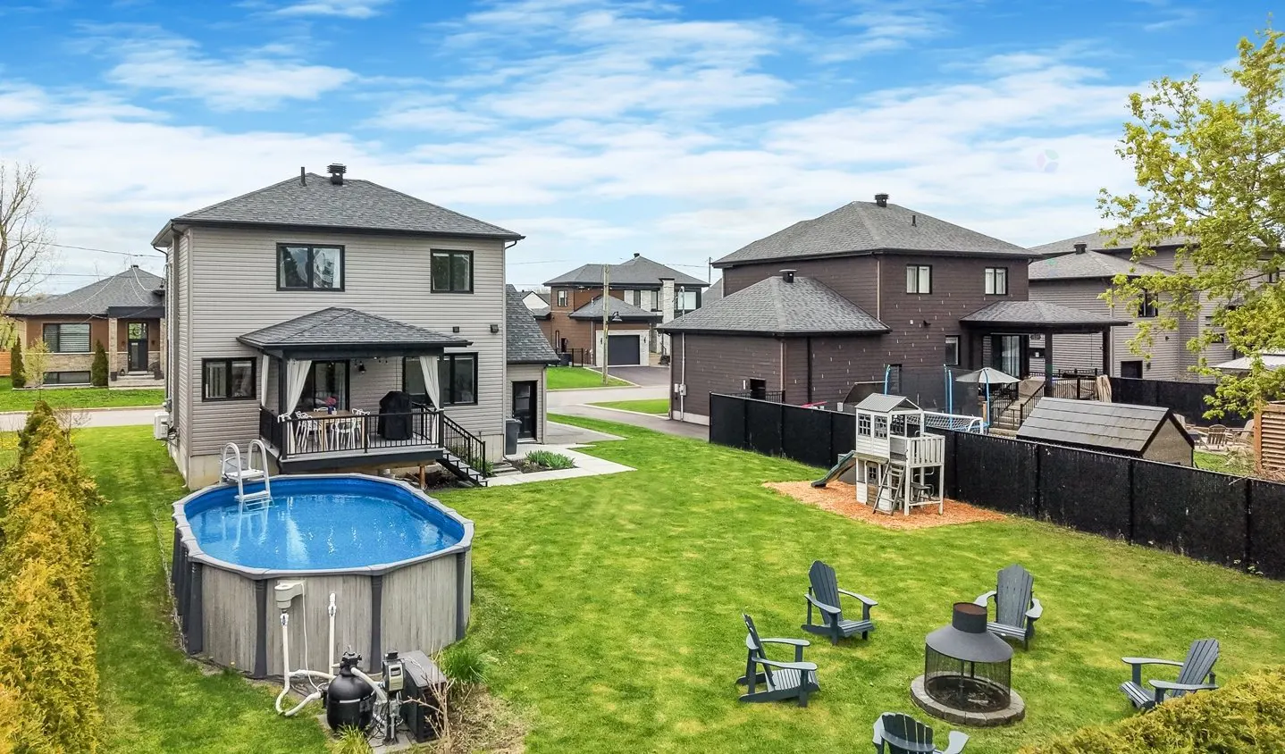 Backyard view of a two-story house with a pool, deck, fire pit, and playground on a green lawn.