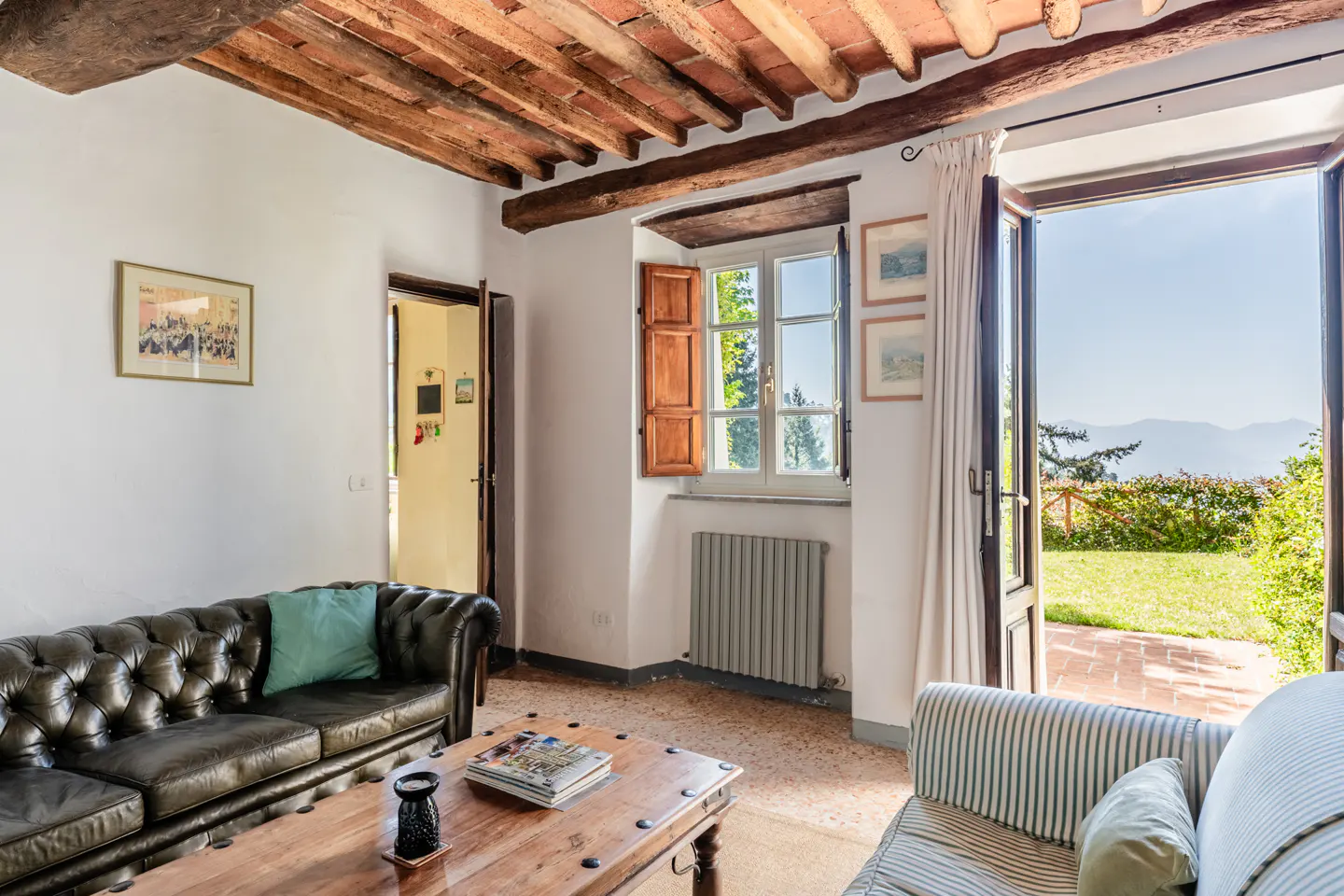 Living room with exposed wood beam ceiling, leather sofa, wood table, and open door to a green lawn and mountain view.