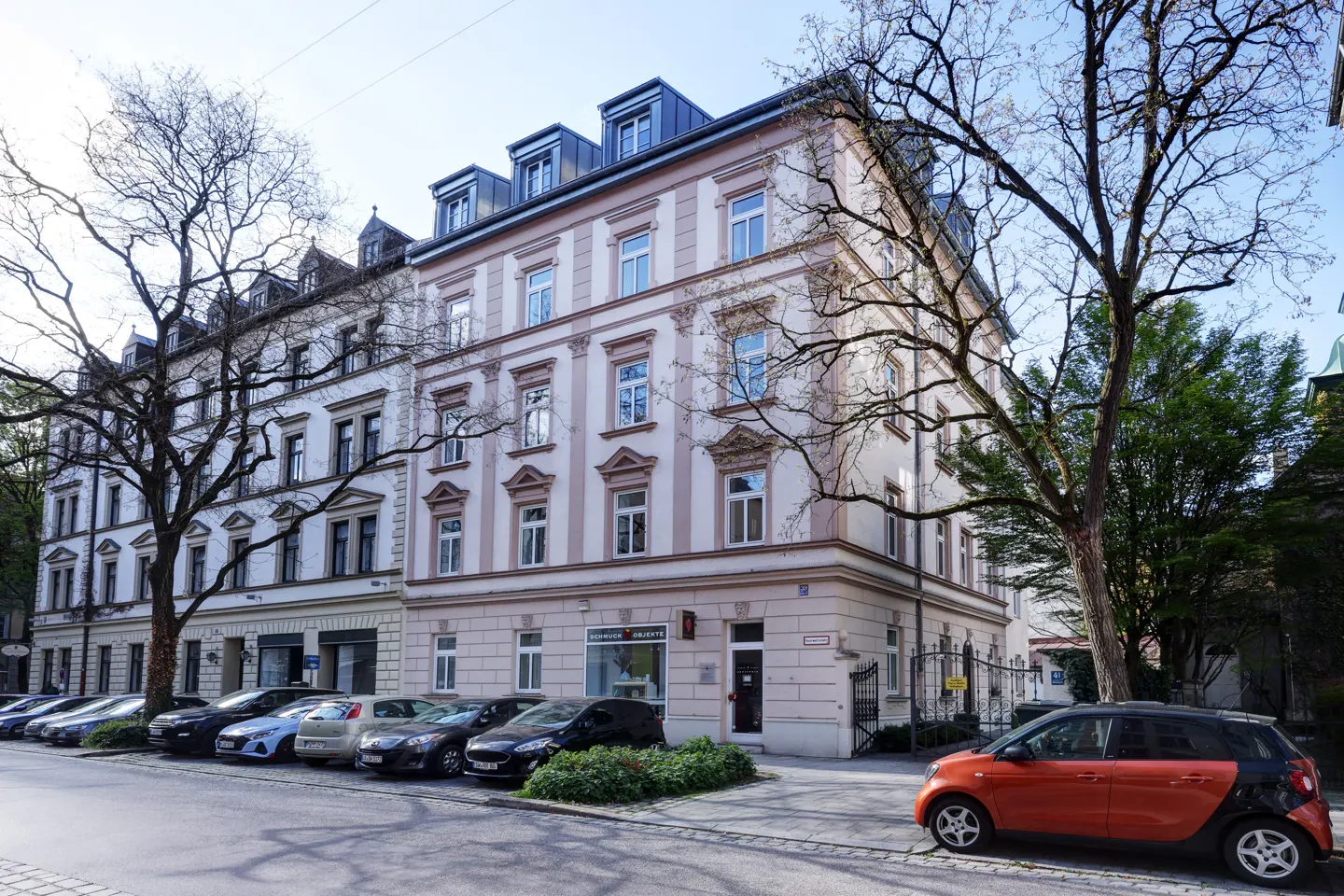 Street view of a multi-story apartment building with parked cars and an orange Smart car.