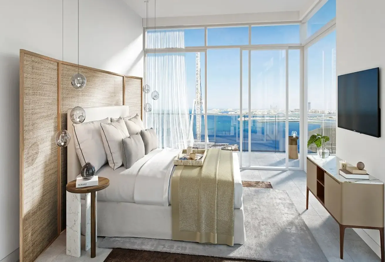 Bright bedroom with a large bed, white linens, and a beige throw blanket. Floor-to-ceiling windows offer a view of the ocean.