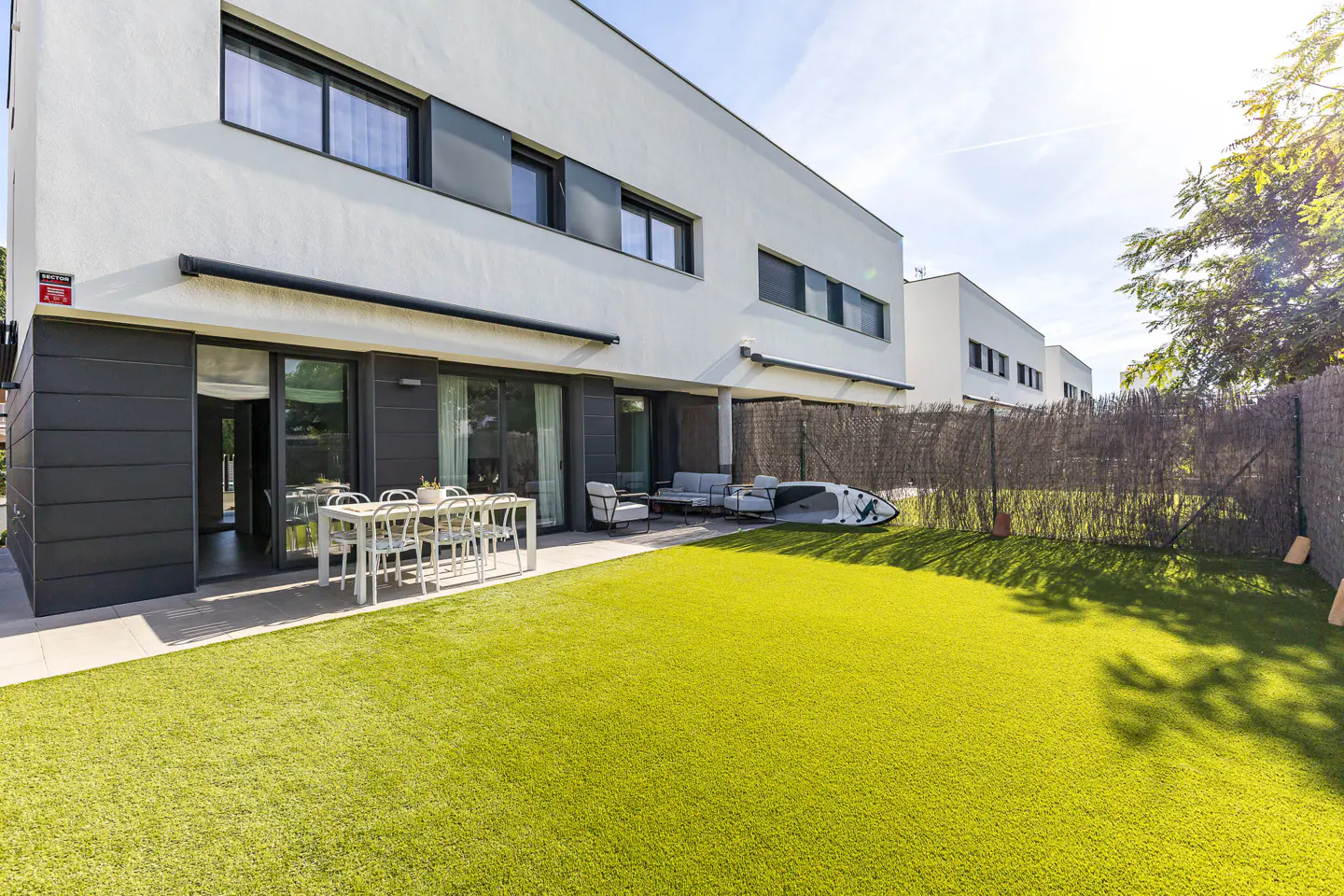 Modern white house with a green lawn, patio furniture, and a paddleboard by a fence.