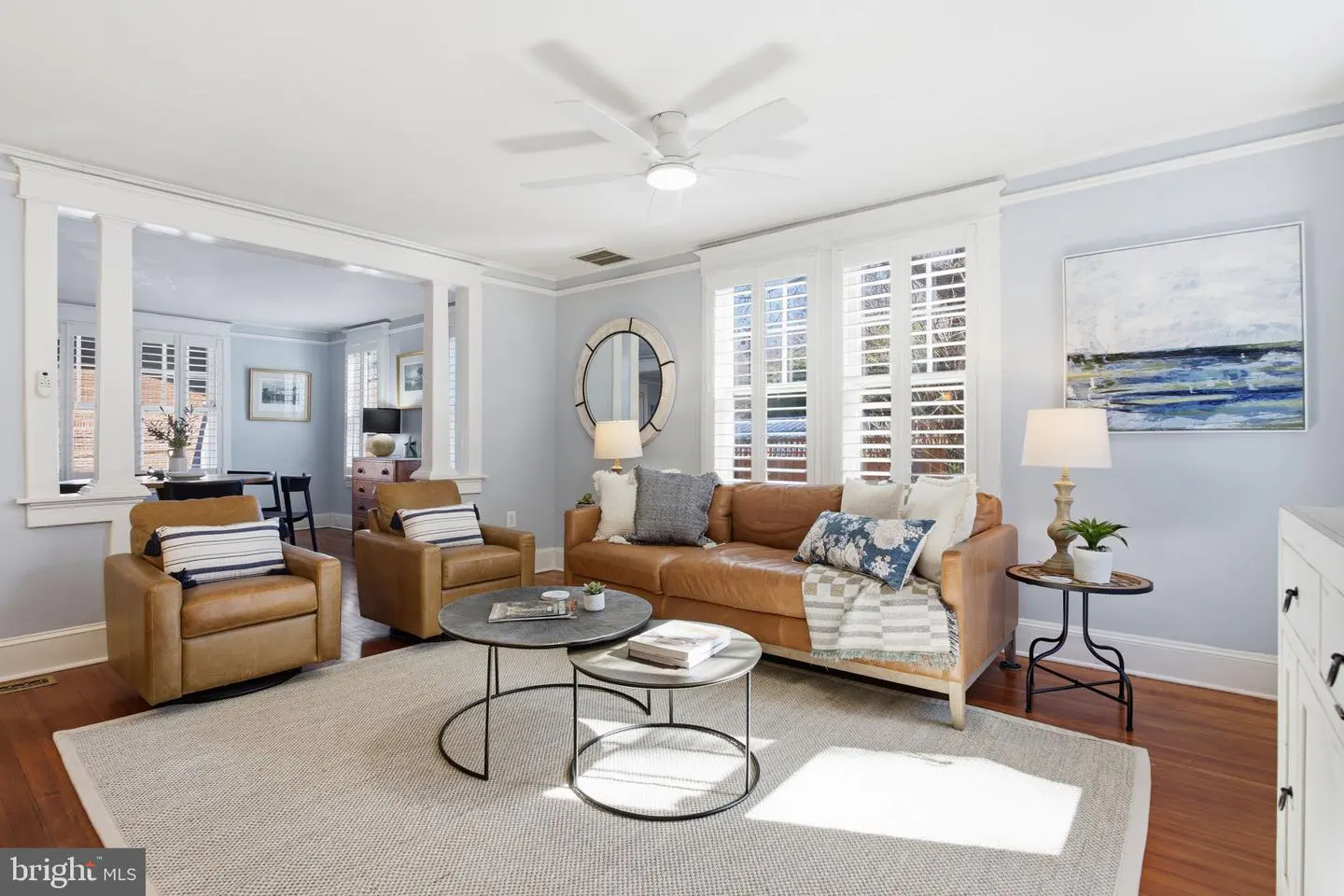 Living room with tan leather sofa and chairs, round nesting tables, and white shutters. A white ceiling fan is overhead.
