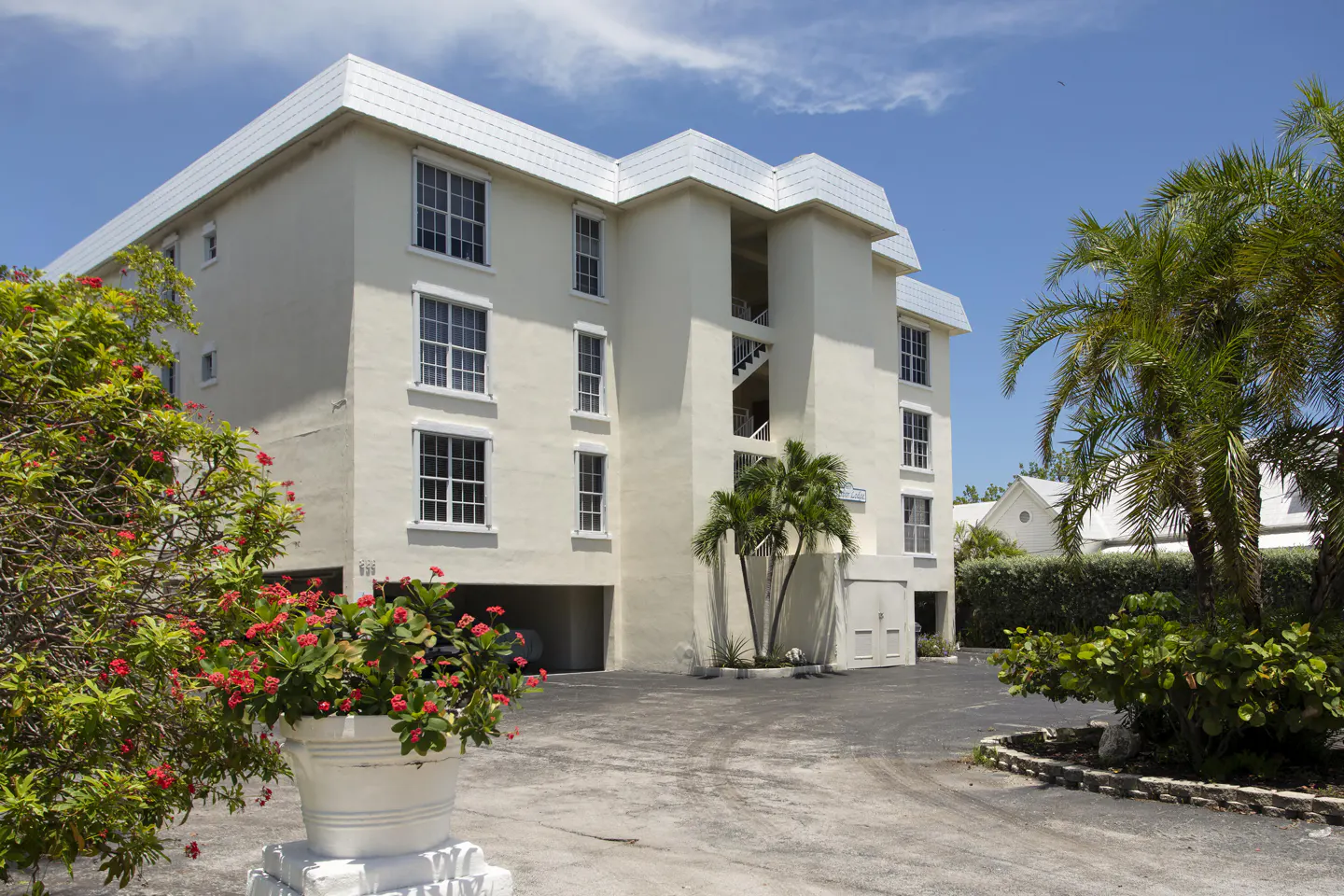 Three-story white apartment building with open stairwell, palm trees, and red flowers in a white pot. Blue sky.