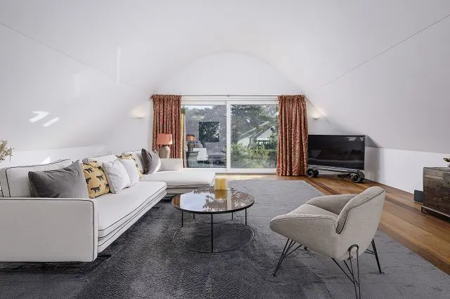 Bright living room with white walls, a white sofa with pillows, a glass coffee table, and a gray chair on a gray rug. A large window with patterned curtains is in the background.