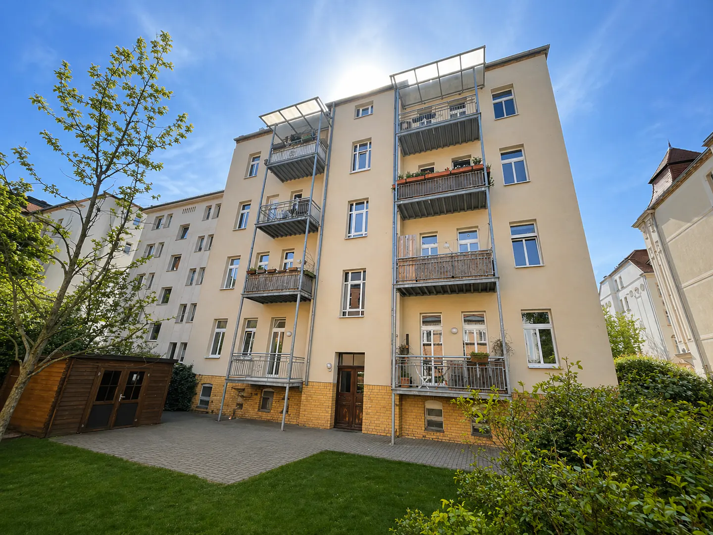 Low-angle view of a beige apartment building with metal balconies, a small wooden shed, and a green lawn under a blue sky.