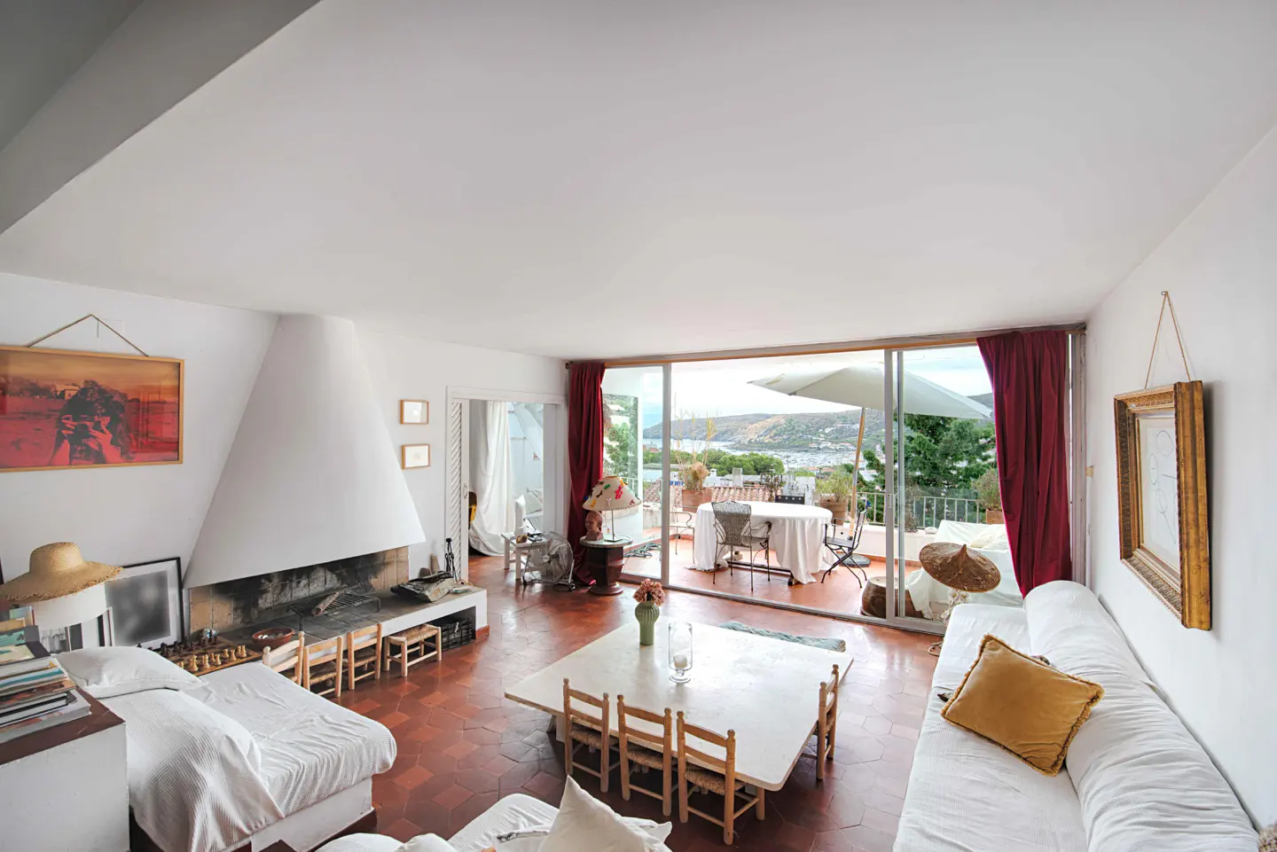 Bright living room with white walls, terracotta floors, a large fireplace, dining area, and balcony with sea views.