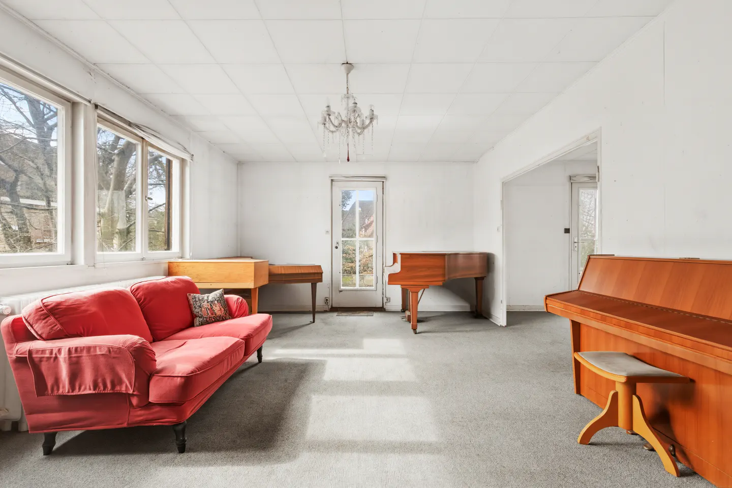 Bright room with a red sofa, three pianos, and a chandelier. Sunlight streams through the windows.