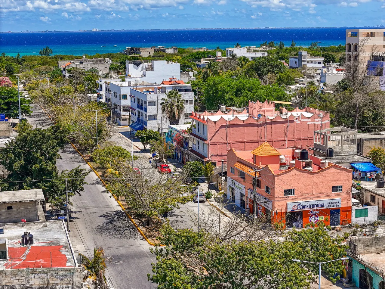 Aerial view of a coastal town with colorful buildings, trees, and the ocean in the background under a blue sky.