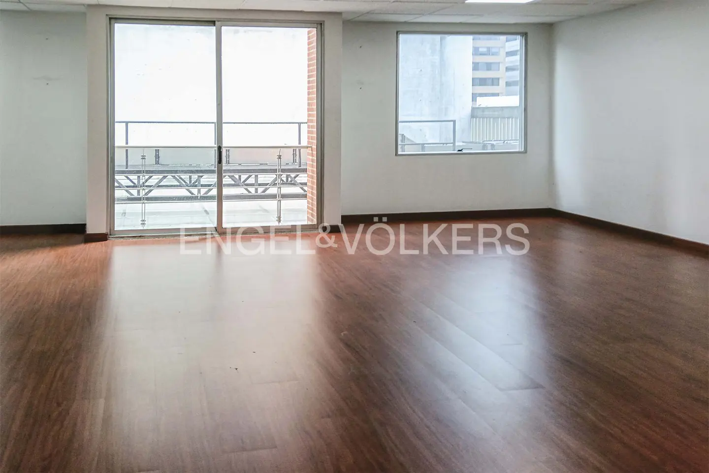 Empty office space with white walls, dark wood-look flooring, a sliding glass door to a balcony, and a large window.