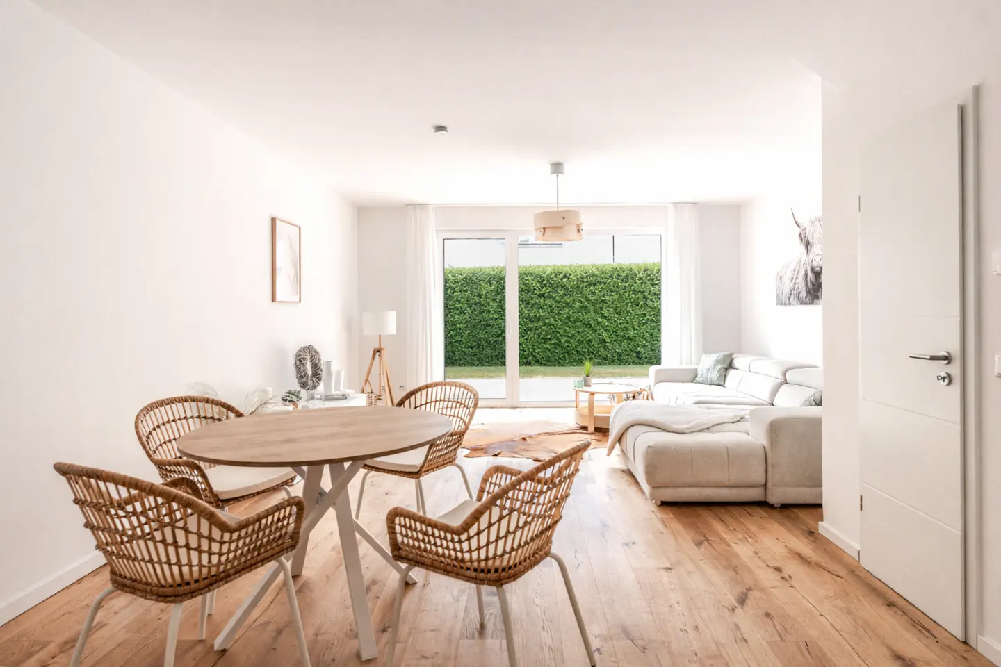 Bright, open-plan living space with wood floors, a round table with wicker chairs, and a white sofa. A green hedge is visible through the glass doors.