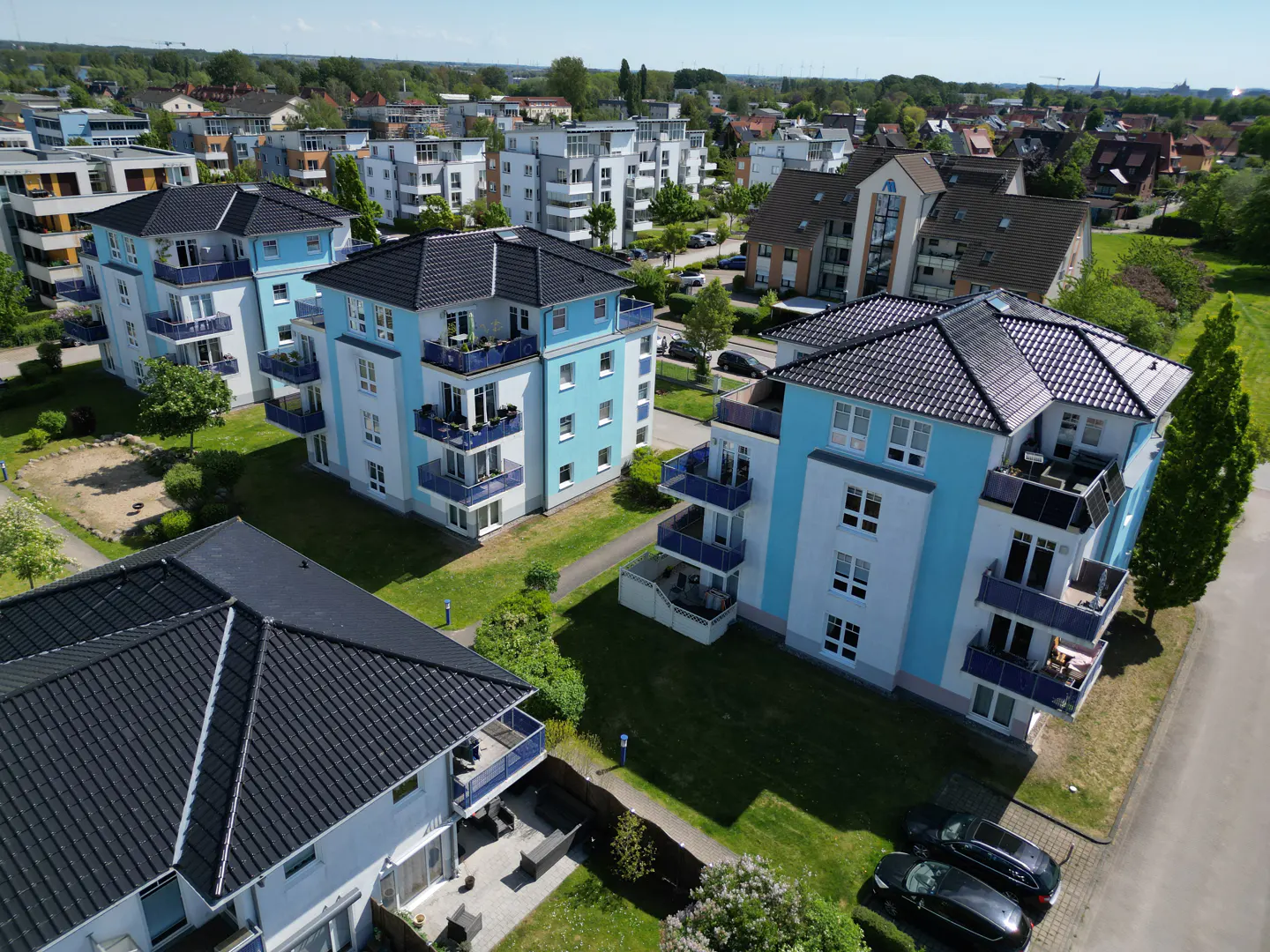 Aerial view of modern light blue and white apartment buildings with dark roofs, balconies, and surrounding green lawns.