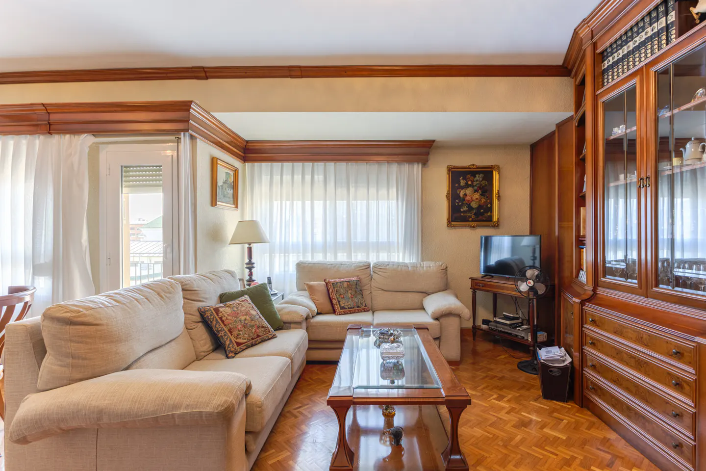 Living room with beige sofas, wood floors, and a glass-topped coffee table. A large wooden bookcase stands against the wall.