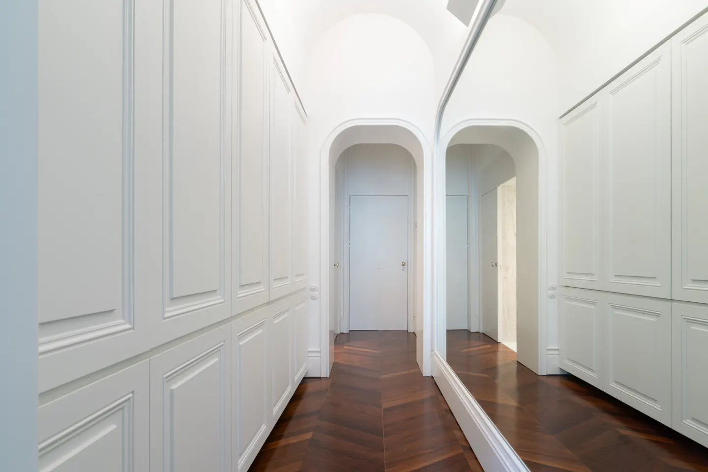 Hallway with white walls, arched doorway, and herringbone wood floor. White cabinets line the left wall, and a mirror reflects the hallway on the right.