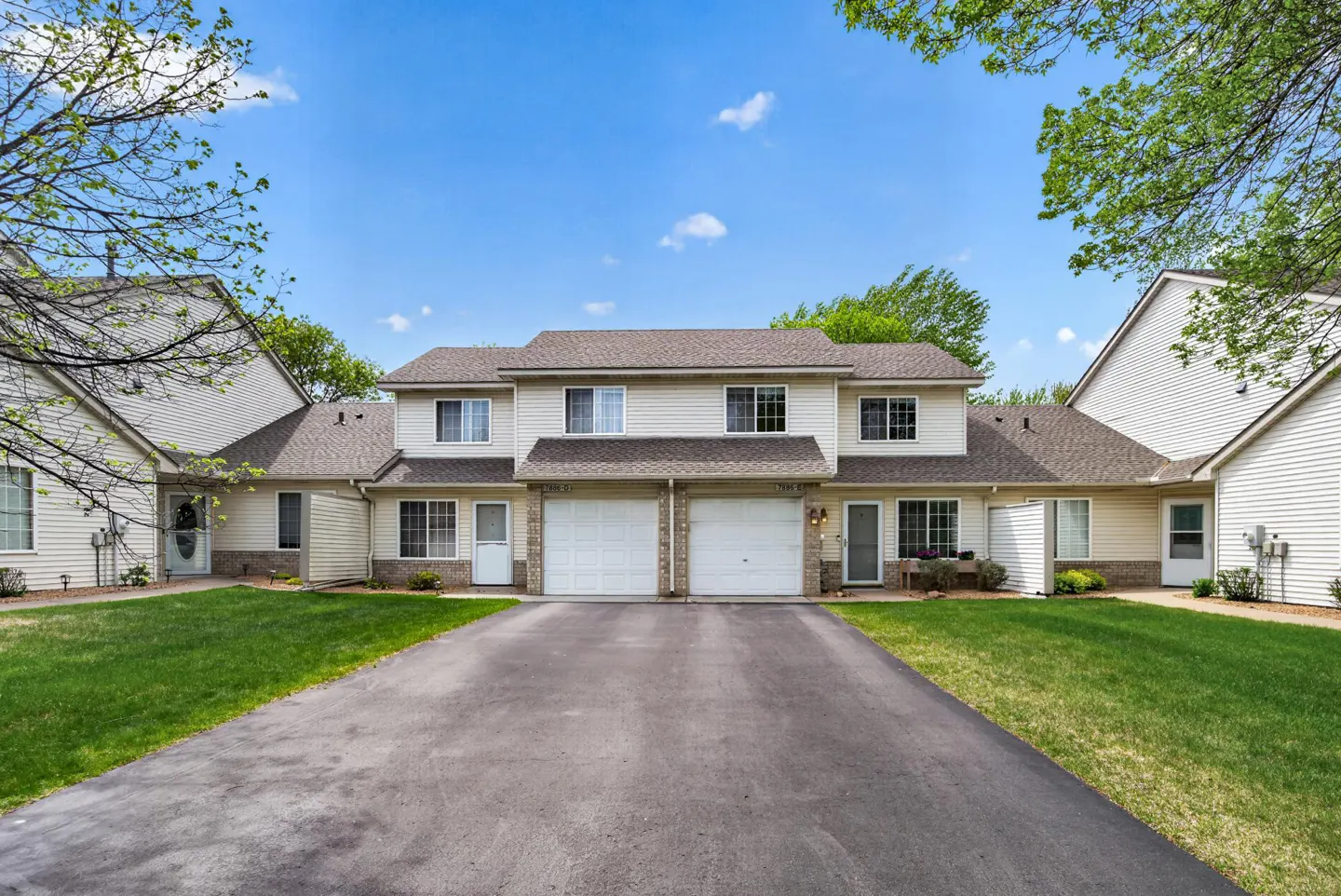 Two-story townhouse with white siding, brown roof, and two-car garage. Green lawn and asphalt driveway. Blue sky and trees in background.