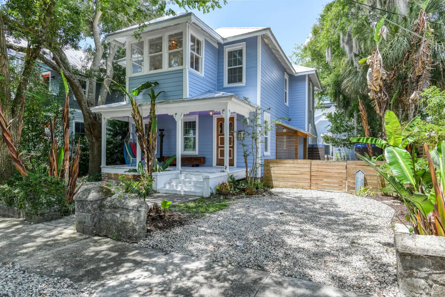 Two-story light blue house with a white porch, white trim, and a gravel driveway surrounded by lush greenery.
