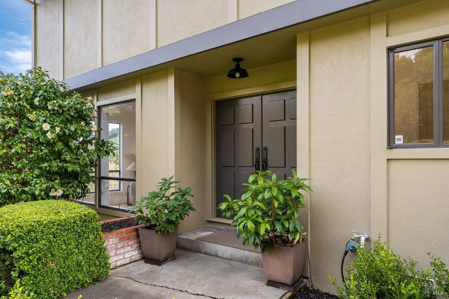 Beige home exterior with a dark double door entrance, potted plants, and trimmed bushes.
