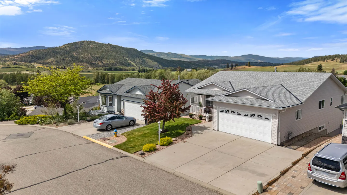 Two-story houses with gray roofs and white garages, cars parked in driveways, and mountains in the background.