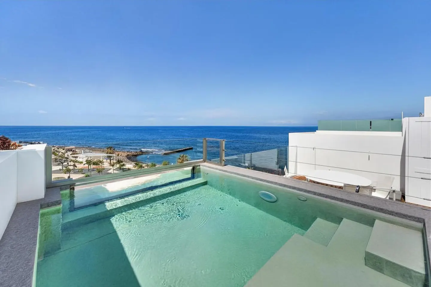 Rooftop pool with ocean view. Turquoise water, gray stone trim, and white building. Blue sky above.
