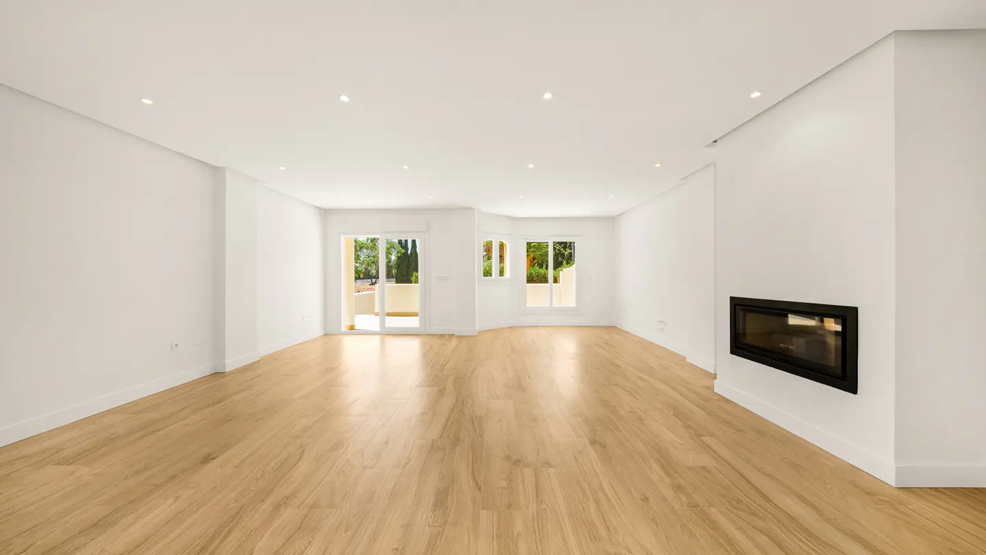 Bright, empty living room with light wood floors, white walls, and recessed lighting. A black fireplace is on the right wall, and sliding glass doors lead to a balcony.