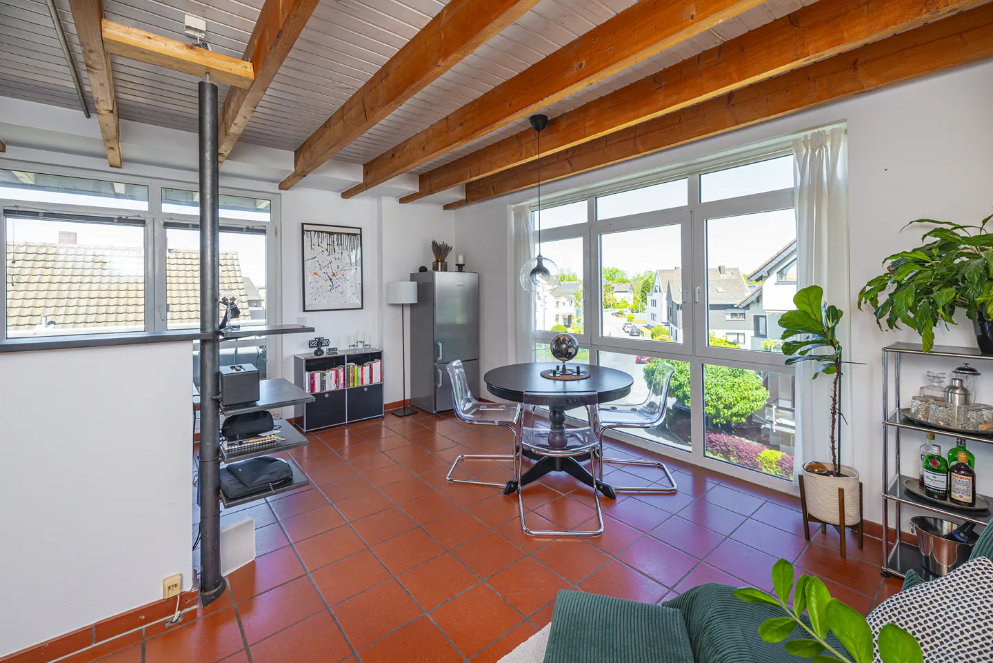 Bright living room with red tile floor, exposed wood beams, and large windows. A black table with clear chairs sits near the window. Shelving unit and plants are also visible.