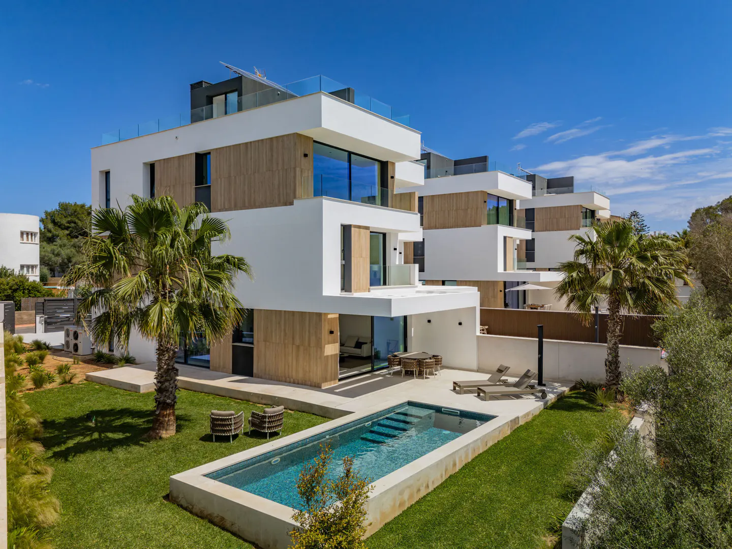 Modern white townhouses with wood accents, a blue pool, and green lawn under a clear blue sky. Palm trees add a tropical feel.