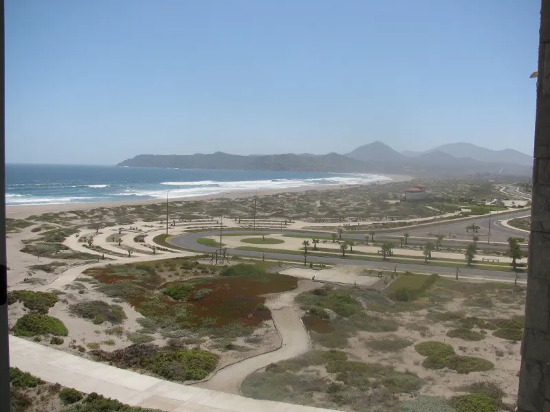 Ocean view from a high rise. Blue water meets sandy beach, with dunes and mountains in the distance. Roads and vegetation in the foreground.
