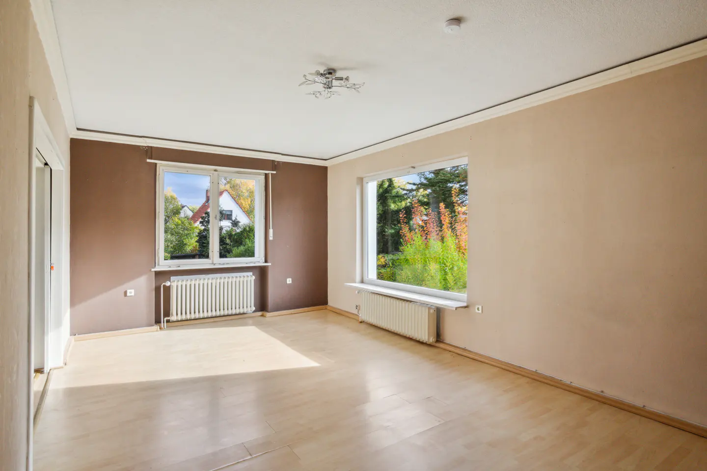 Empty room with light wood floors, beige and brown walls, and two windows showing trees and a house.