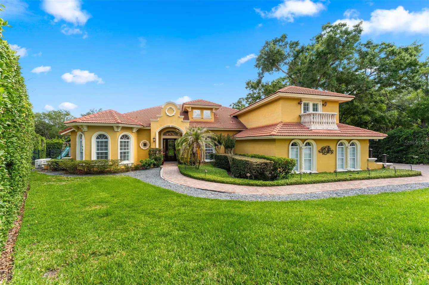 Two-story yellow house with a red tile roof, arched windows, and a balcony. A green lawn and trees surround the house.