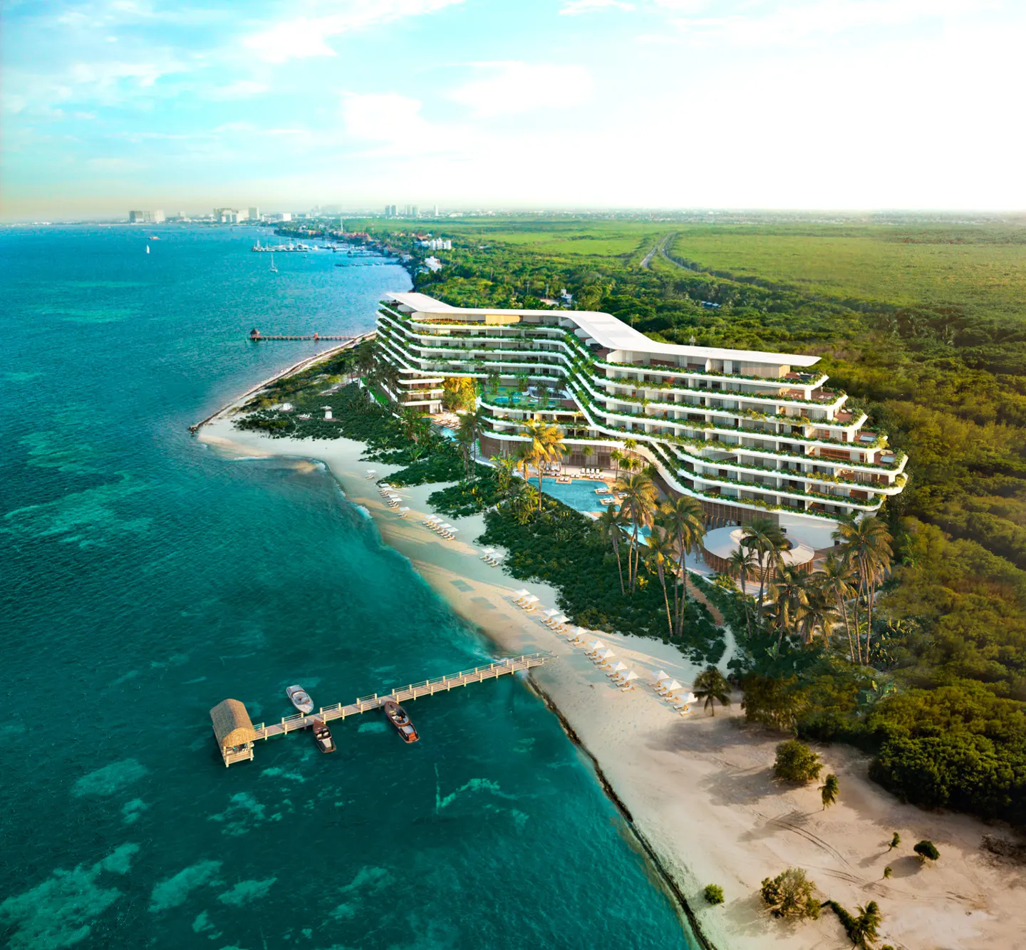 Aerial view of a modern, white beachfront resort with a pier extending into turquoise water. Lush green vegetation surrounds the building.