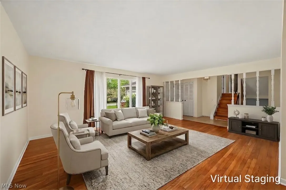 A staged living room with hardwood floors, a gray rug, a sofa, two chairs, and a wooden coffee table. Stairs are visible in the background.