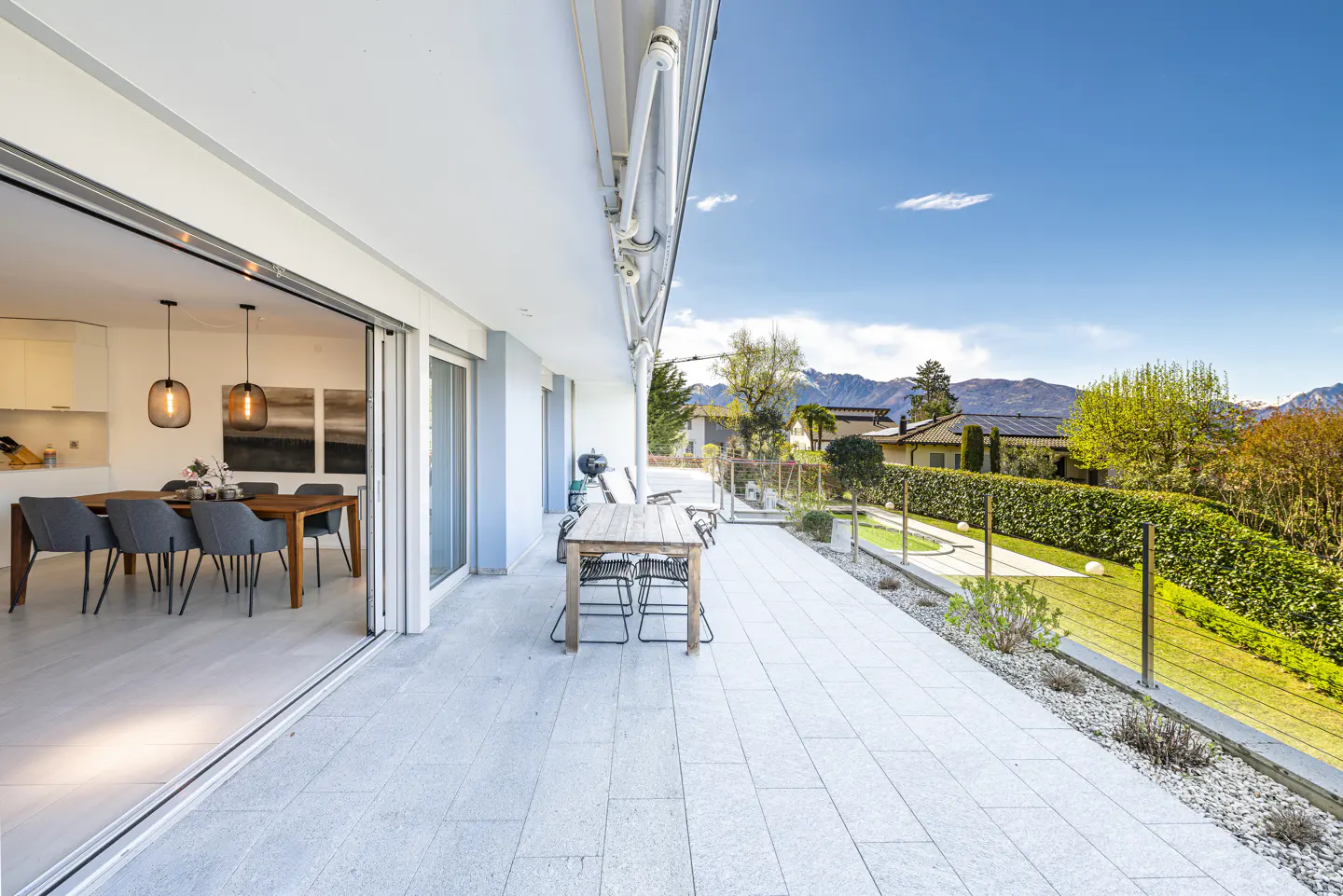 Bright, modern patio with dining table and chairs, open to a dining room. Mountain view in the background under a blue sky.