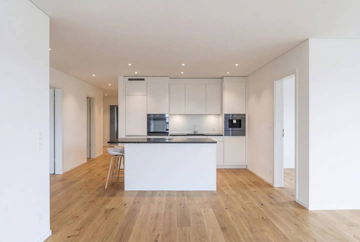 Bright, modern kitchen with white cabinets, black countertops, and a central island with stools. Light wood floors and white walls create a clean, spacious feel.