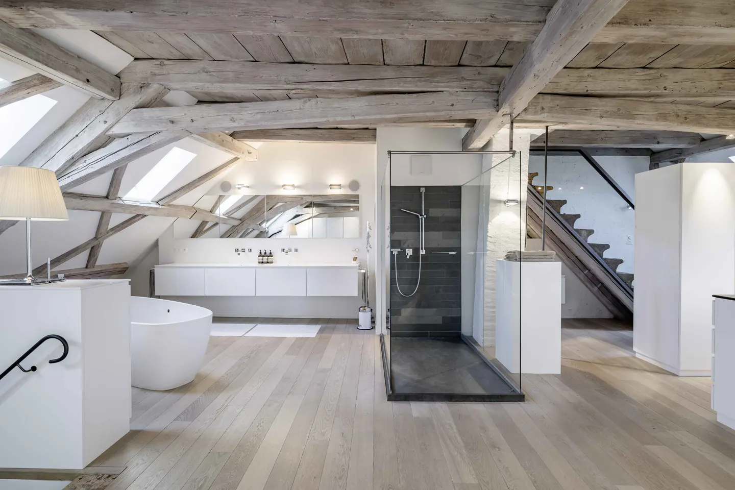 Attic bathroom with exposed wood beams, skylights, and light wood floors. Features a white tub, vanity, and glass shower.