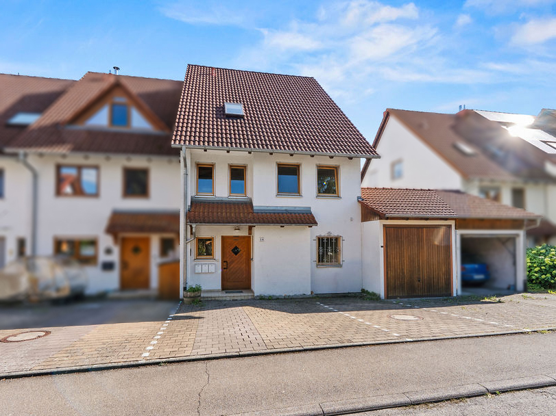 White two-story house with a brown tiled roof, wooden front door, and attached garage, set on a paved driveway. [AI-generated text]