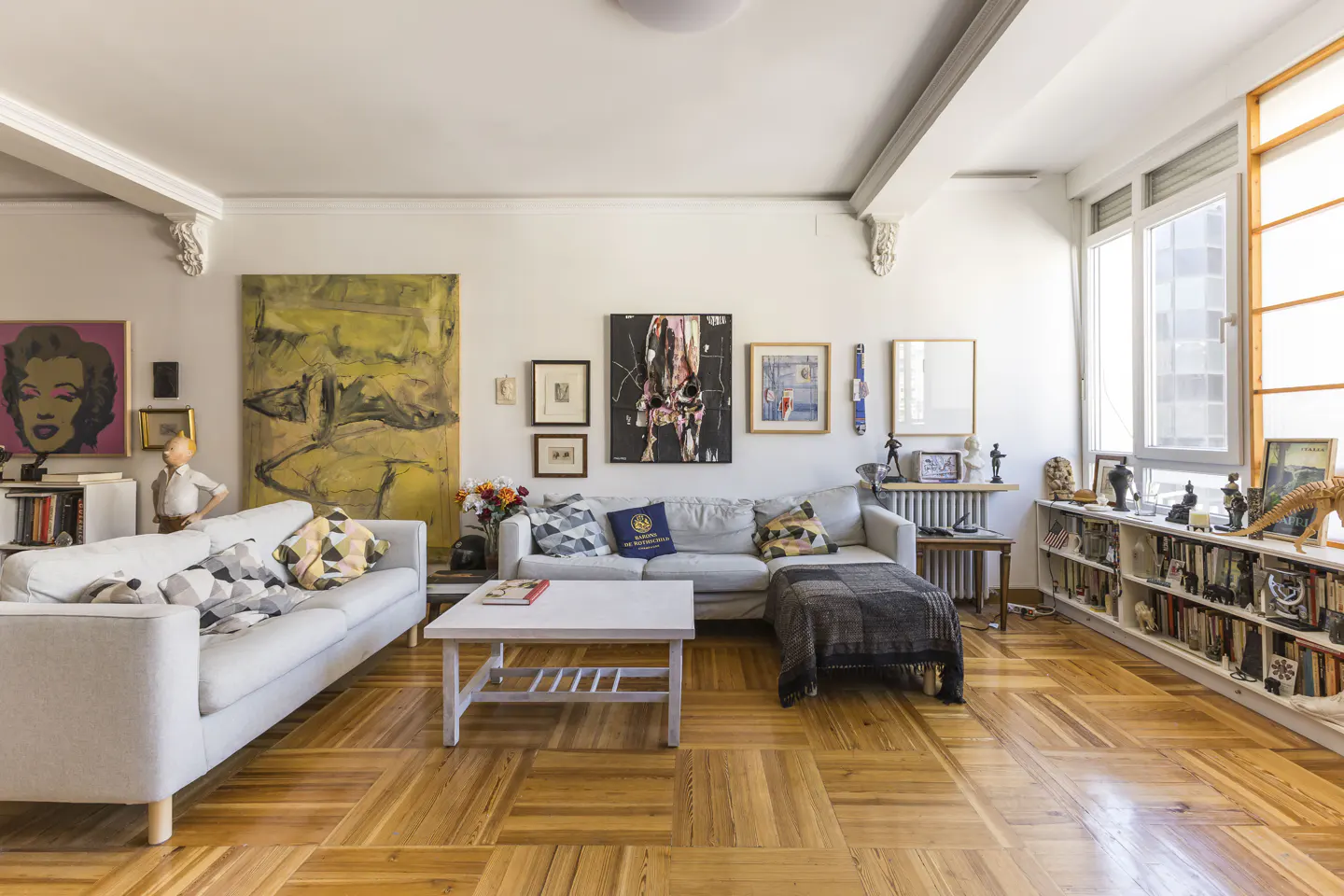 Bright living room with parquet floors, white walls, and large windows. Two sofas, a white coffee table, and a bookshelf filled with books and decorative objects.