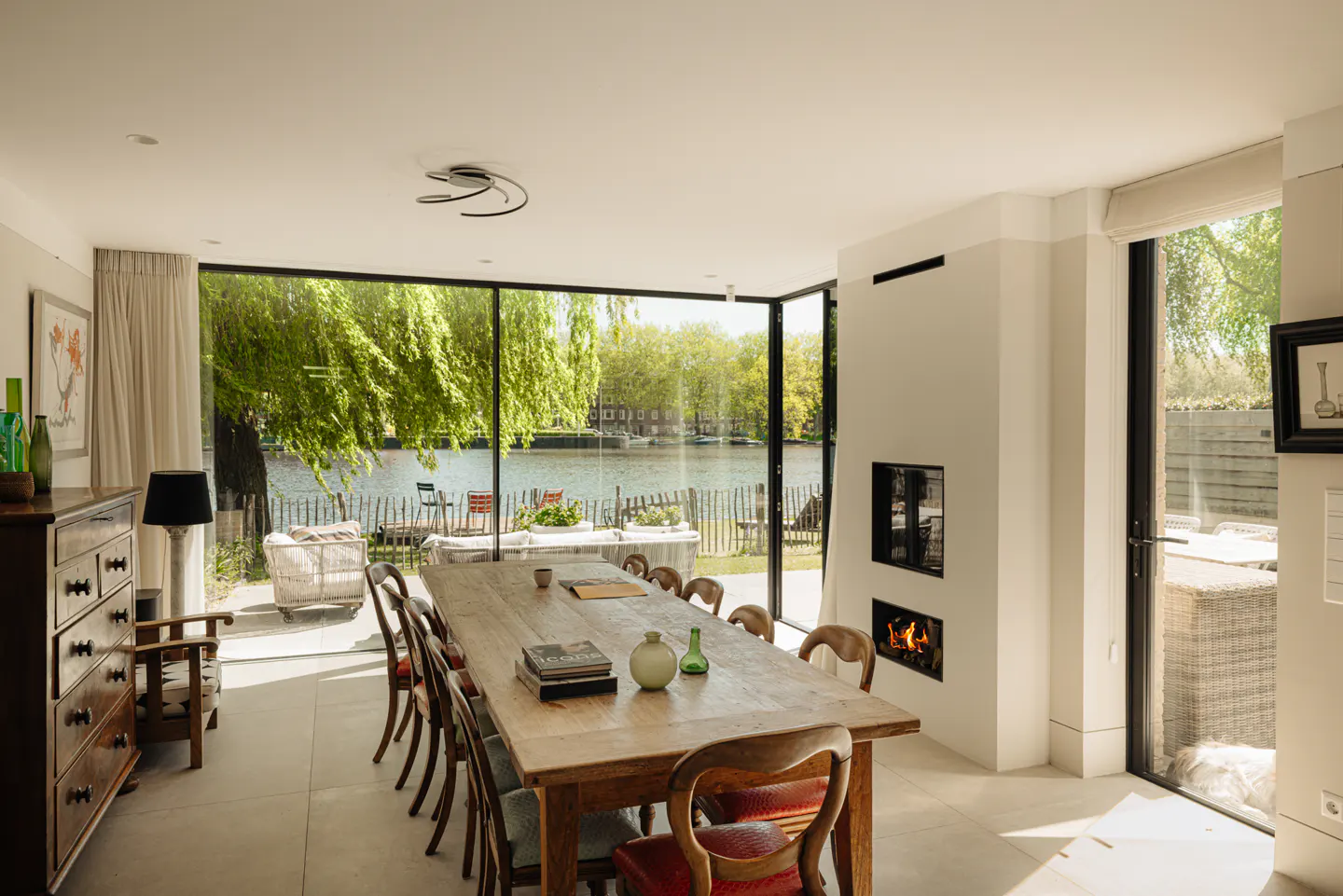 Bright dining room with a long wooden table, chairs, and a fireplace. Large windows offer a view of a river and greenery.