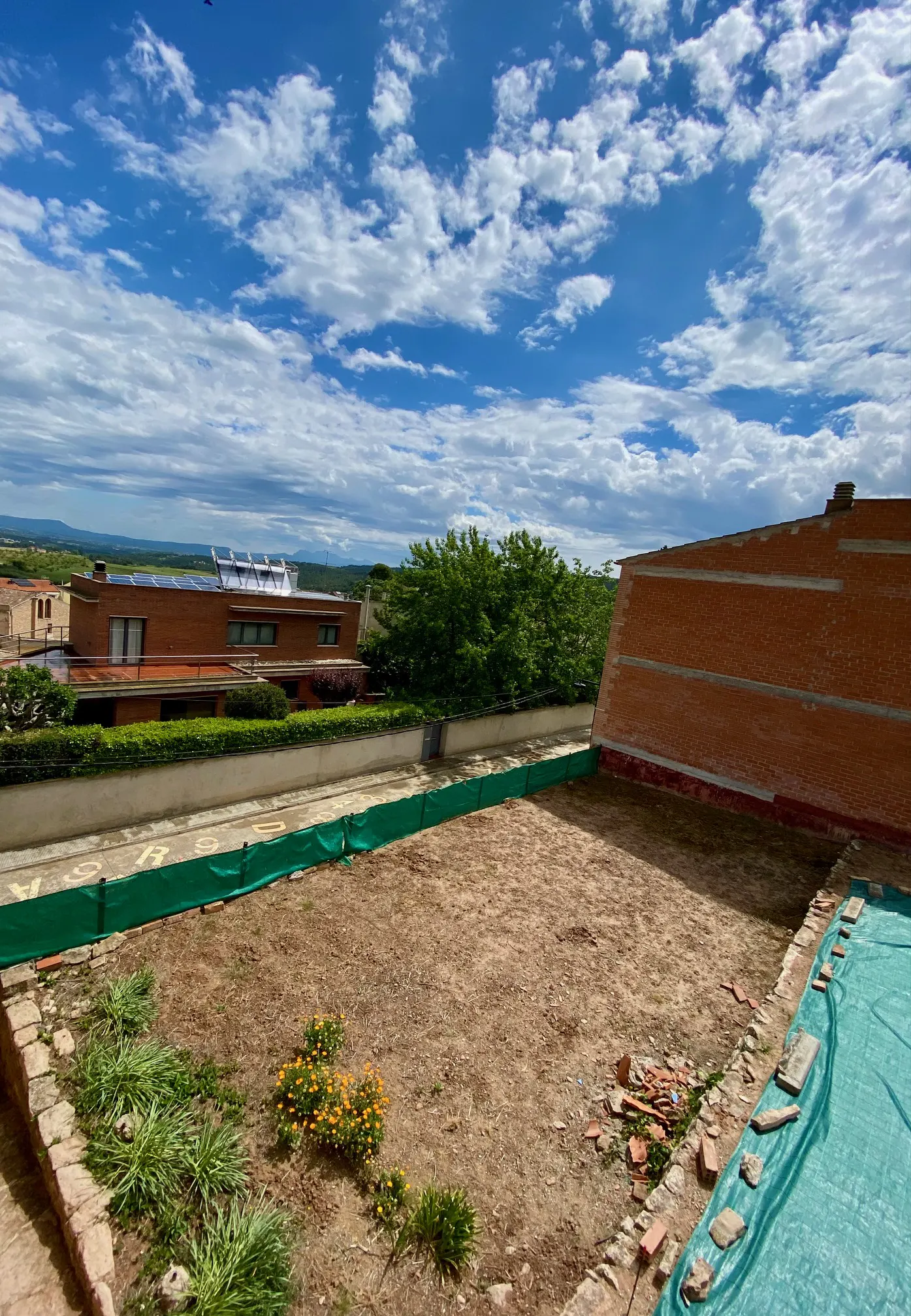 View of a property's empty lot with a brick wall, green fence, and a house in the background under a cloudy blue sky.