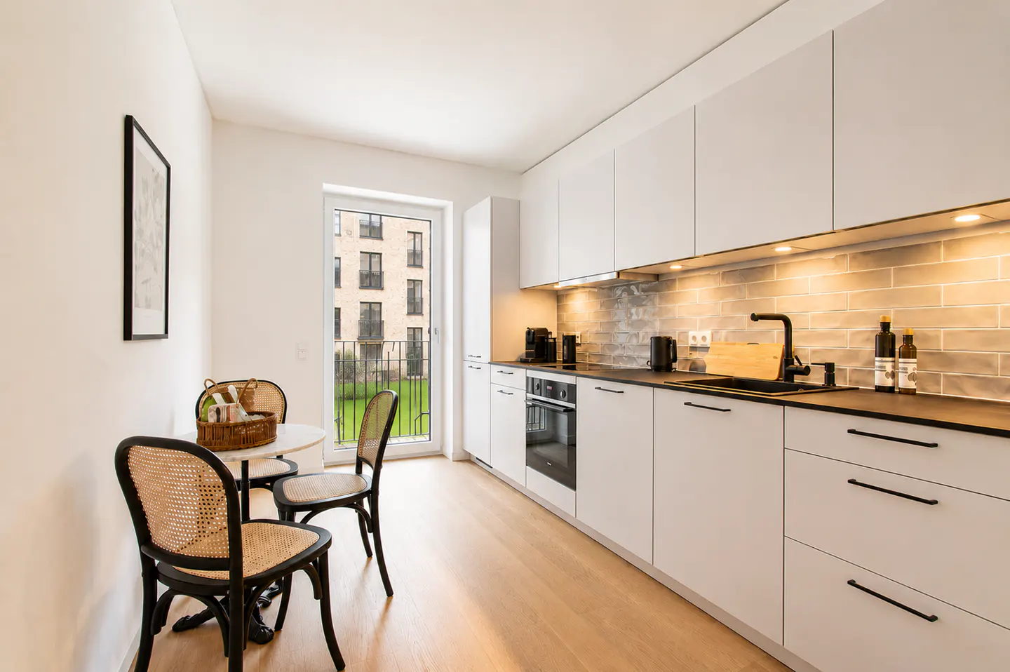 Modern kitchen with white cabinets, black hardware, light wood floors, and a small dining area with two rattan chairs.