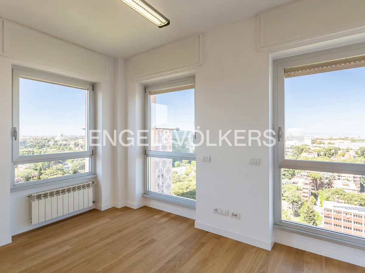 Bright, empty room with wood floors, white walls, and three large windows offering city views. A radiator sits beneath one window.