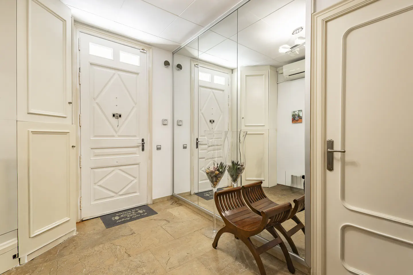 Entryway with a white door, mirrored wall, and two wooden chairs. A welcome mat sits on the stone floor.